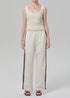 A person stands against a plain background wearing an off white Citizens Of Humanity/AGOLDE aurora track pant with a brown stripe, a cream ribbed tank top, black sandals, a gold necklace, and a bracelet. Their face is not shown.