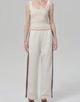 A person stands against a plain background wearing an off white Citizens Of Humanity/AGOLDE aurora track pant with a brown stripe, a cream ribbed tank top, black sandals, a gold necklace, and a bracelet. Their face is not shown.