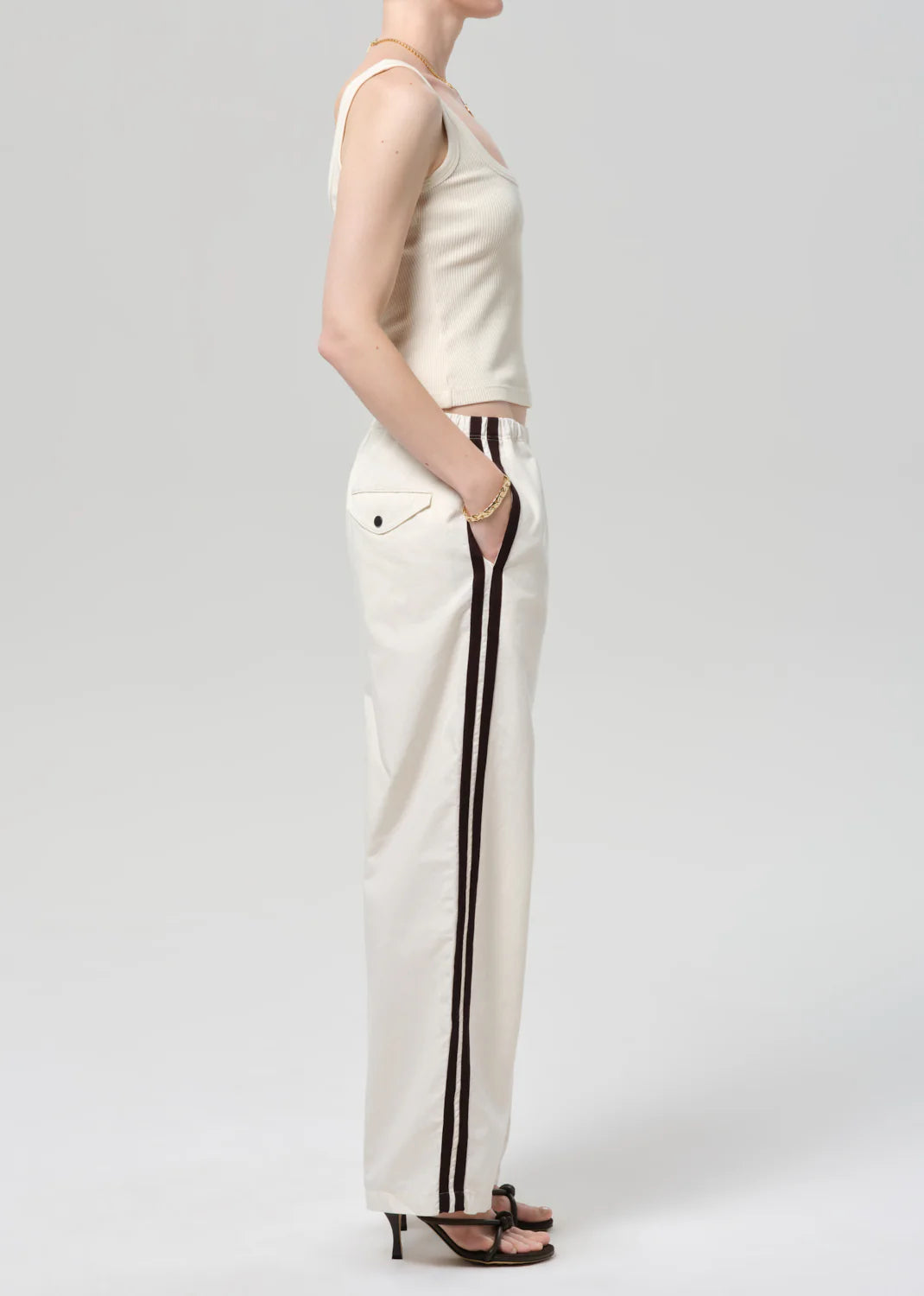 A person stands in profile, hands in pockets, wearing a sleeveless beige top, Citizens Of Humanity/AGOLDE aurora track pant in off white with dark side stripes, and black heeled sandals against a plain light background.