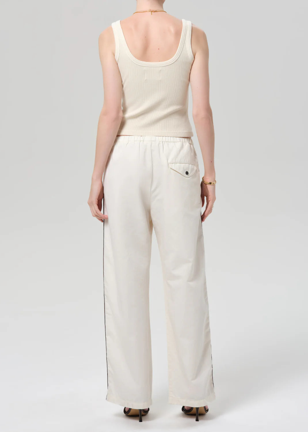 A person faces away from the camera, wearing Citizens Of Humanity/AGOLDE's aurora track pant in off white with black side piping and a back pocket, made from an Italian cotton blend, paired with a sleeveless ribbed white tank top.
