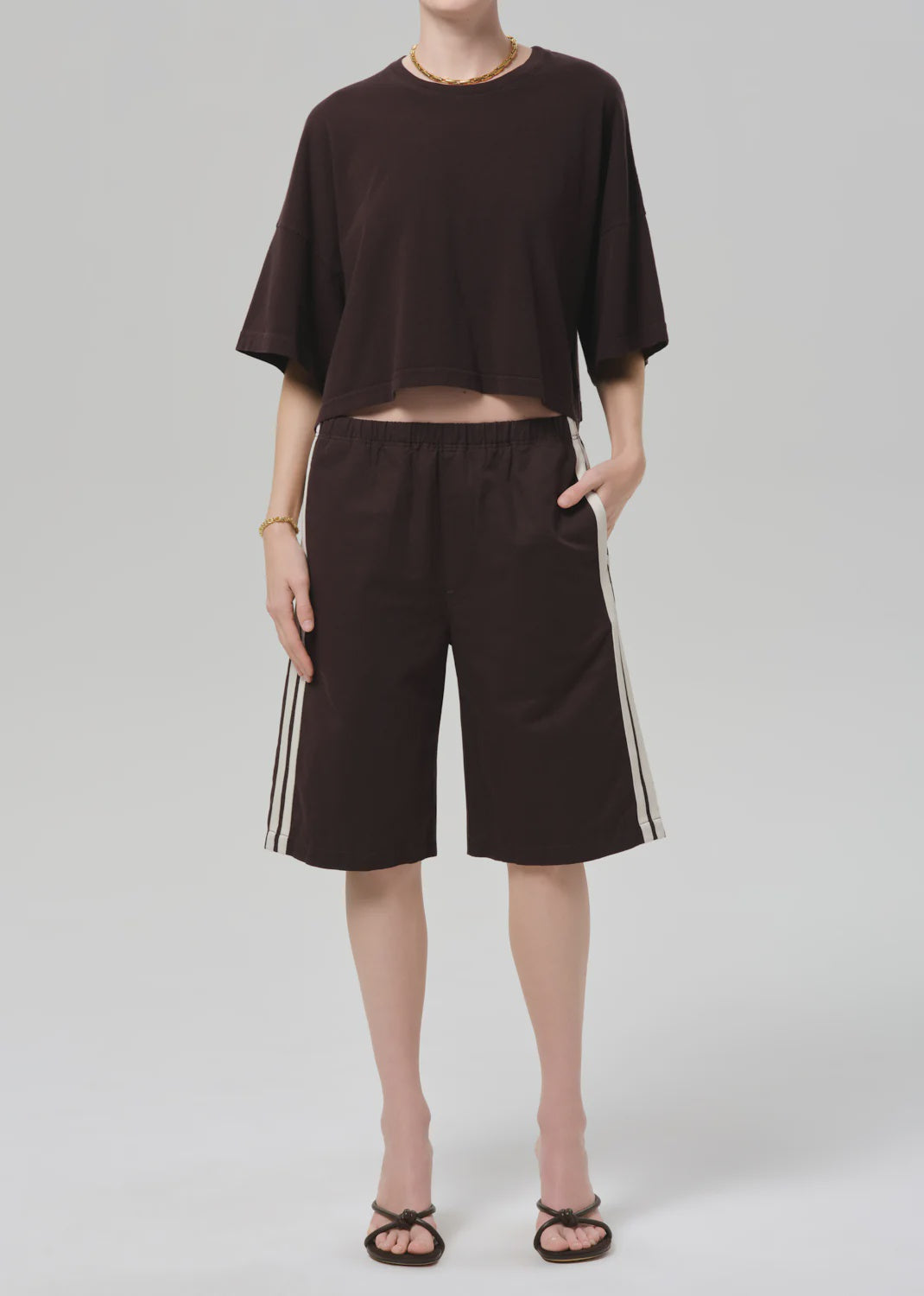 Wearing a loose dark brown Italian cotton blend cropped T-shirt, Citizens Of Humanity/AGOLDE anais pull on track shorts with white side stripes, a gold necklace and bracelet, and black sandals, a person stands against a plain light gray background.
