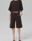 Wearing a loose dark brown Italian cotton blend cropped T-shirt, Citizens Of Humanity/AGOLDE anais pull on track shorts with white side stripes, a gold necklace and bracelet, and black sandals, a person stands against a plain light gray background.