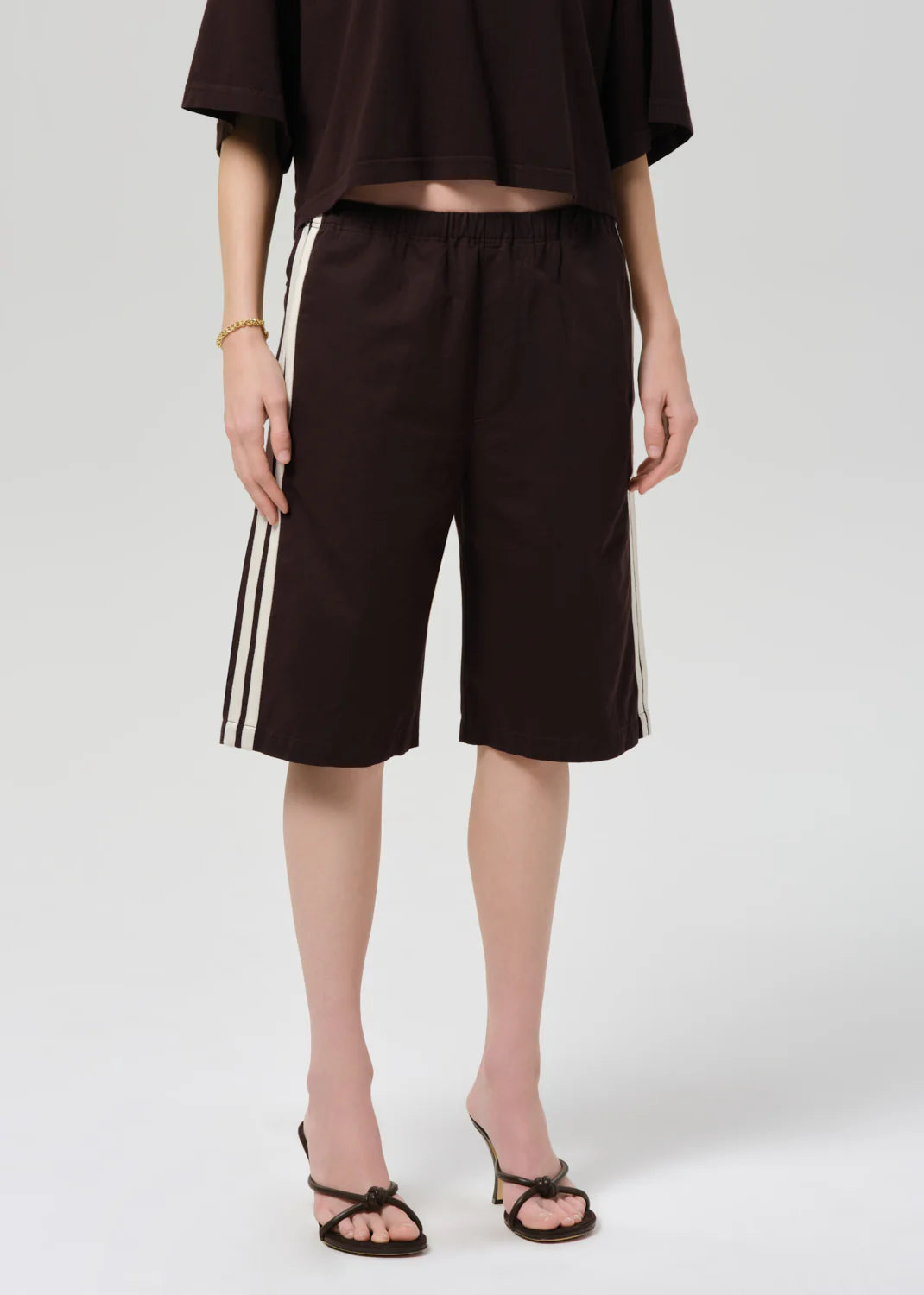 A person wears Citizens Of Humanity/AGOLDE anais pull on track shorts—loose, dark brown, longline with white side stripes—paired with a matching oversized Italian cotton-blend top, gold bracelet, and black heeled sandals against a plain light background.