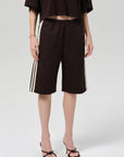 A person wears Citizens Of Humanity/AGOLDE anais pull on track shorts—loose, dark brown, longline with white side stripes—paired with a matching oversized Italian cotton-blend top, gold bracelet, and black heeled sandals against a plain light background.