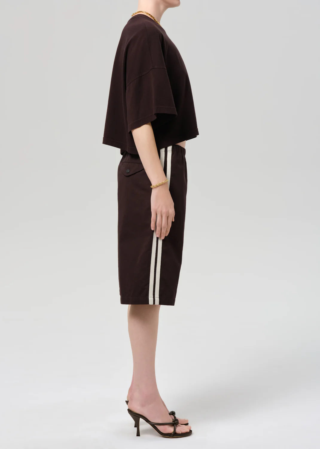 A person stands in profile wearing a loose dark brown T-shirt, Citizens Of Humanity/AGOLDE anais pull on track short—Italian cotton blend with white side stripes—a gold bracelet, a necklace, and black heeled sandals against a plain light grey background.