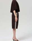 A person stands in profile wearing a loose dark brown T-shirt, Citizens Of Humanity/AGOLDE anais pull on track short—Italian cotton blend with white side stripes—a gold bracelet, a necklace, and black heeled sandals against a plain light grey background.