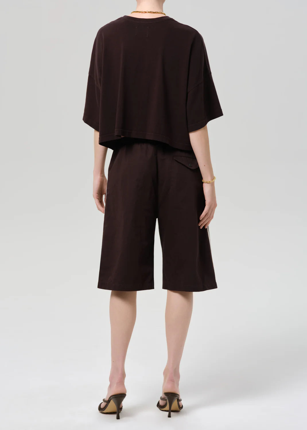 Facing away, a person wears a loose dark brown Italian cotton blend short-sleeve top and matching Citizens Of Humanity/AGOLDE anais pull on track shorts, styled with heeled sandals and a bracelet against a plain light background.