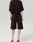 Facing away, a person wears a loose dark brown Italian cotton blend short-sleeve top and matching Citizens Of Humanity/AGOLDE anais pull on track shorts, styled with heeled sandals and a bracelet against a plain light background.