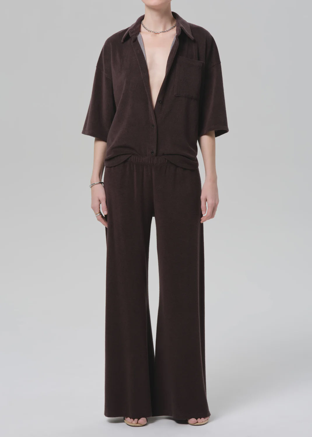 A person models the Citizens Of Humanity/AGOLDE nora pull on pant—a loose, dark brown, wide-leg style—paired with a matching short-sleeve button-up shirt, standing barefoot against a plain light gray background.