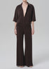 A person models the Citizens Of Humanity/AGOLDE nora pull on pant—a loose, dark brown, wide-leg style—paired with a matching short-sleeve button-up shirt, standing barefoot against a plain light gray background.