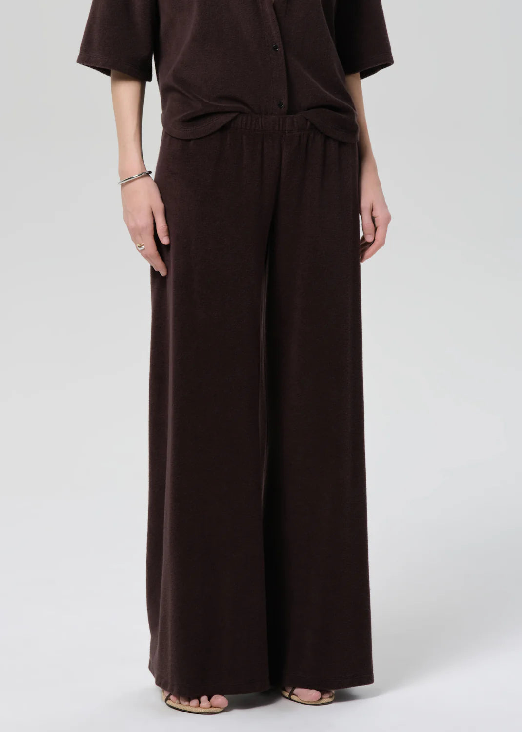 A person wears Citizens Of Humanity/AGOLDE’s nora pull on pant—a loose, wide-leg dark brown style—paired with a matching top. The relaxed lower half and part of the arms appear against a light gray background.