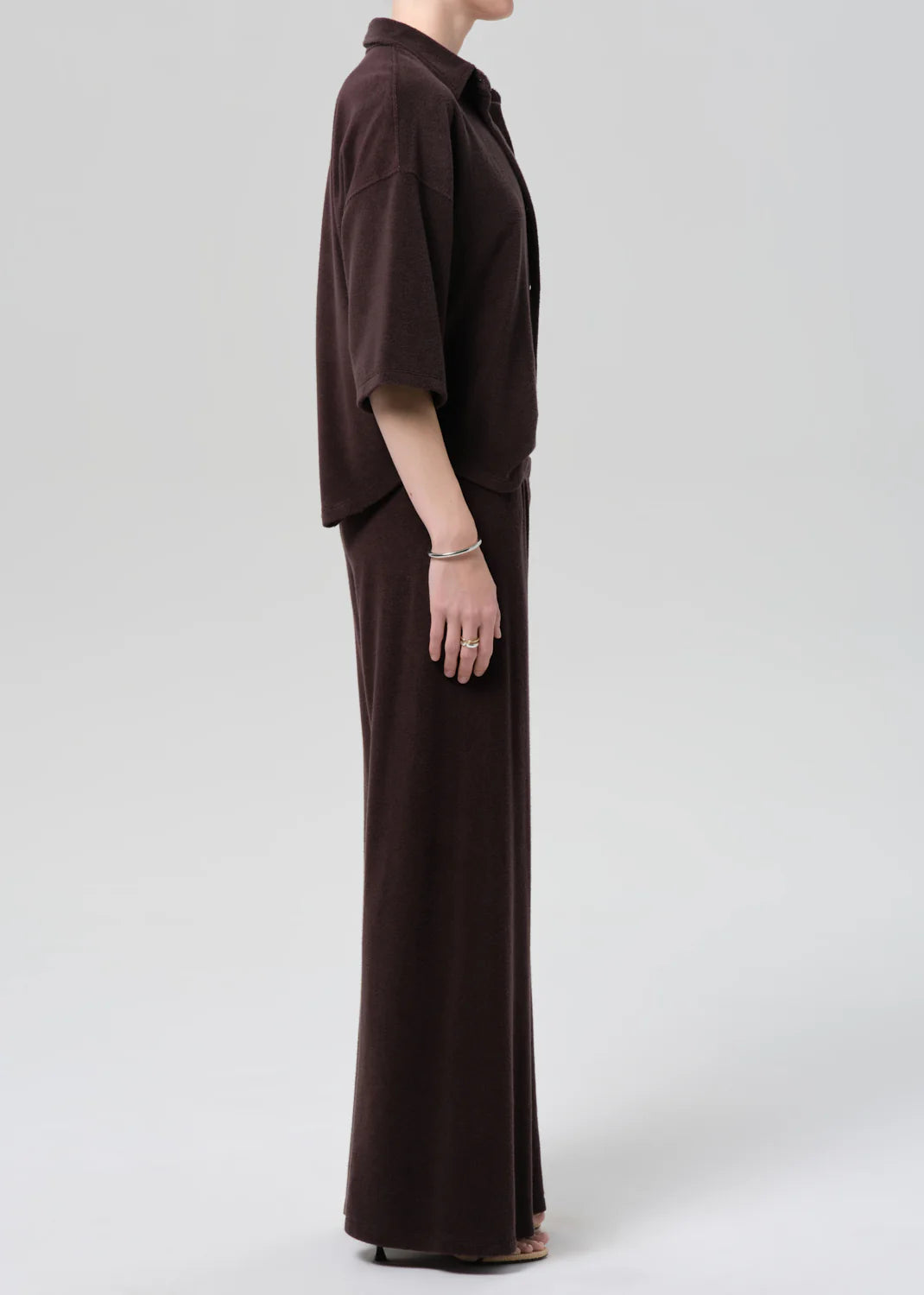 A person stands in profile against a plain background, wearing the Citizens Of Humanity/AGOLDE nora pull on pant in dark brown microterry cotton blend with a loose-fitting top and sandals. The relaxed silhouette appears soft and comfortable.