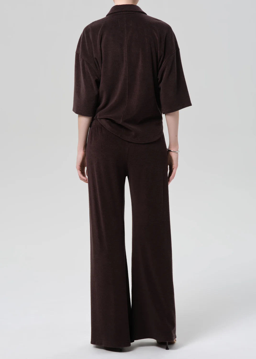 Seen from the back, a person stands against a light gray background wearing Citizens Of Humanity/AGOLDE's nora pull on pant—a wide-leg, dark brown microterry cotton blend set with a loose long-sleeve top. The relaxed fit looks soft and comfortable.
