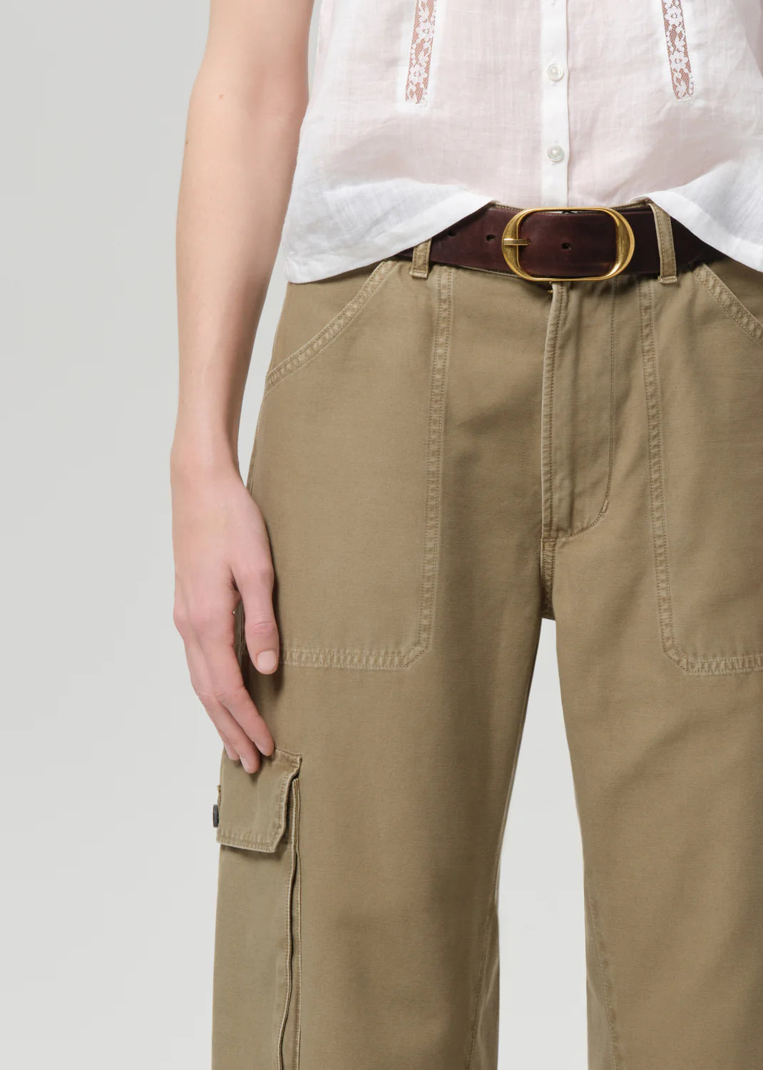A person wears a white short-sleeve button-up shirt tucked into Citizens Of Humanity/AGOLDE thassos twisted seam utility pants with cargo pockets and a dark brown belt with a large gold buckle. Only the torso and upper legs are visible.