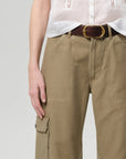 A person wears a white short-sleeve button-up shirt tucked into Citizens Of Humanity/AGOLDE thassos twisted seam utility pants with cargo pockets and a dark brown belt with a large gold buckle. Only the torso and upper legs are visible.