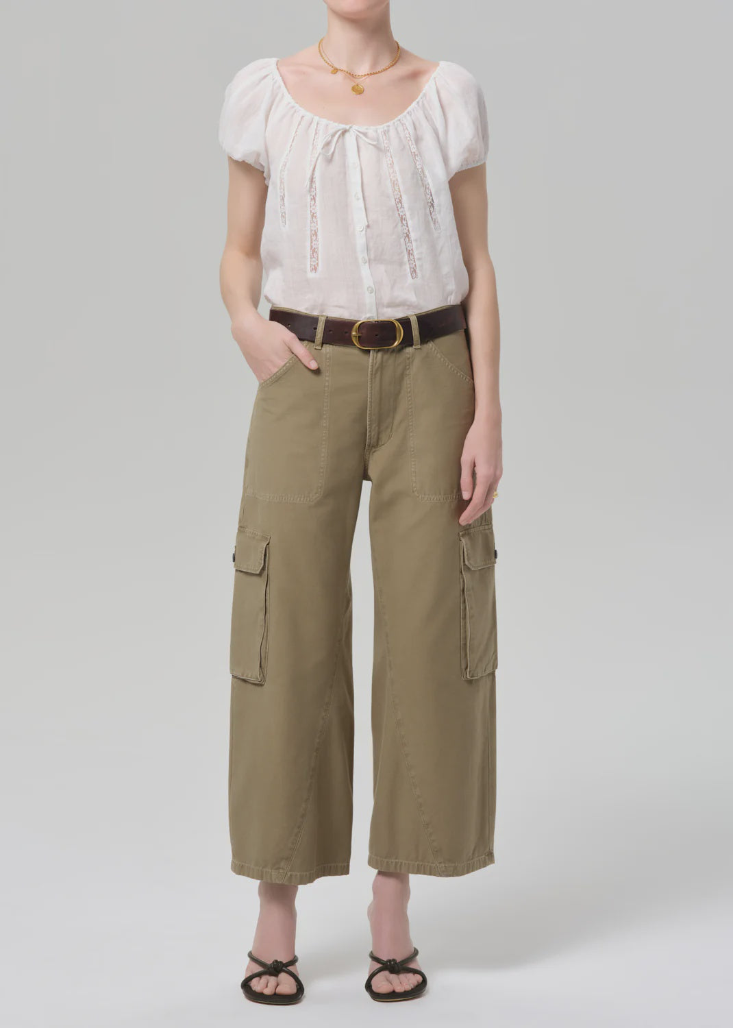 A person wearing the Citizens Of Humanity/AGOLDE thassos twisted seam utility pant in khaki with cargo pockets, a white short-sleeve blouse, brown belt, black strappy sandals, and gold necklace stands against a plain light gray background.