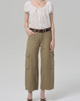 A person wearing the Citizens Of Humanity/AGOLDE thassos twisted seam utility pant in khaki with cargo pockets, a white short-sleeve blouse, brown belt, black strappy sandals, and gold necklace stands against a plain light gray background.