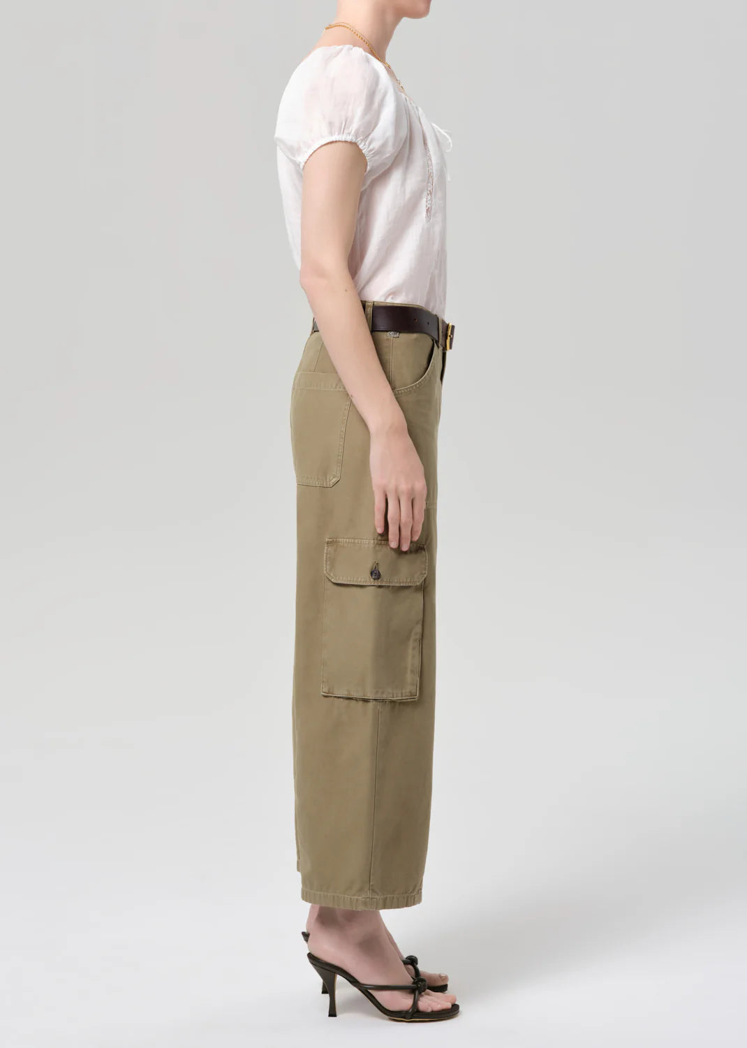 A woman stands in profile against a light gray background wearing Citizens Of Humanity/AGOLDE thassos twisted seam utility pant, a white short-sleeve blouse, dark belt, and black heeled sandals.