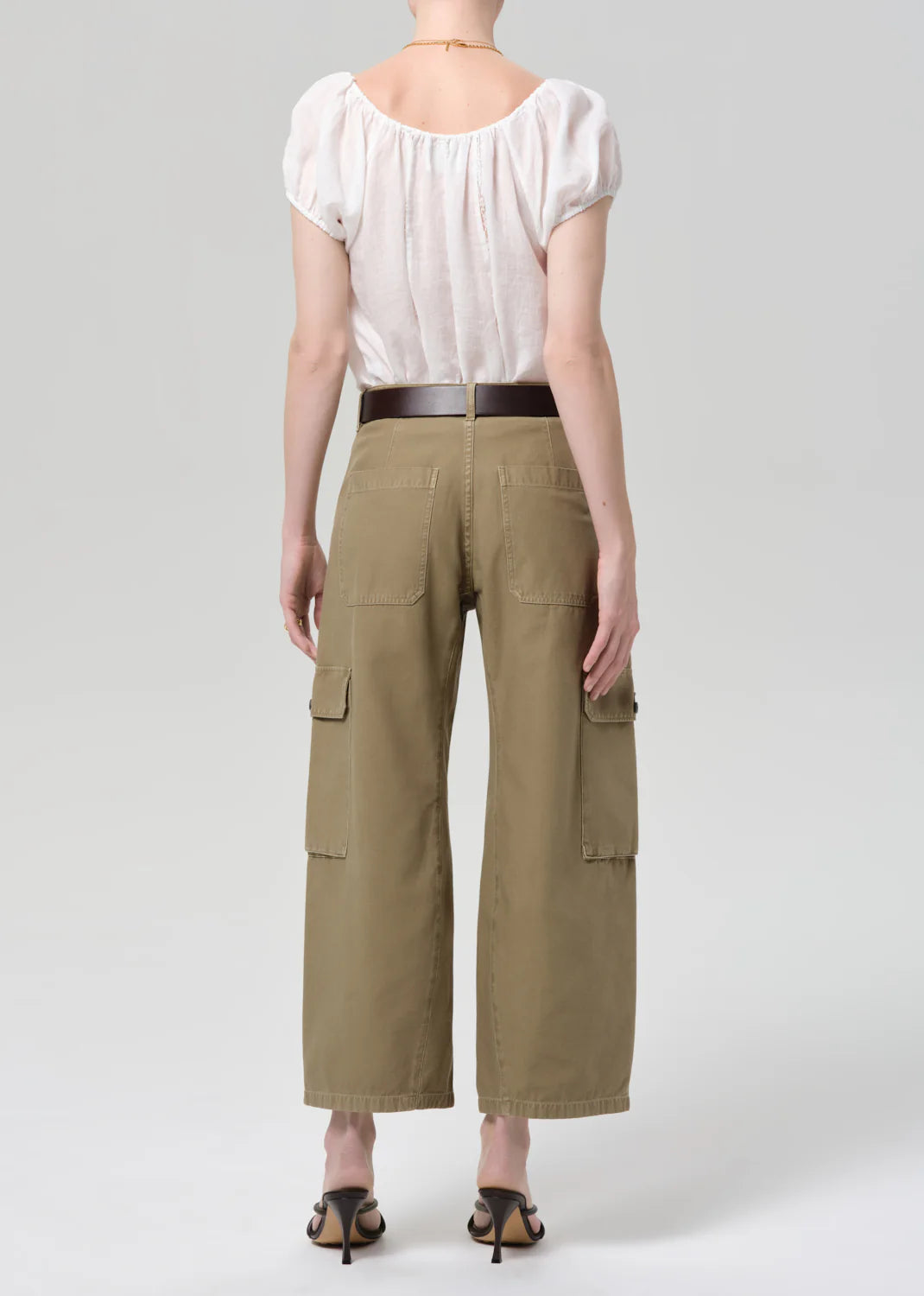 A person stands facing away from the camera, wearing a white short-sleeve blouse and Citizens Of Humanity thassos twisted seam utility pant in khaki, styled with a dark belt and black heeled sandals against a plain light background.