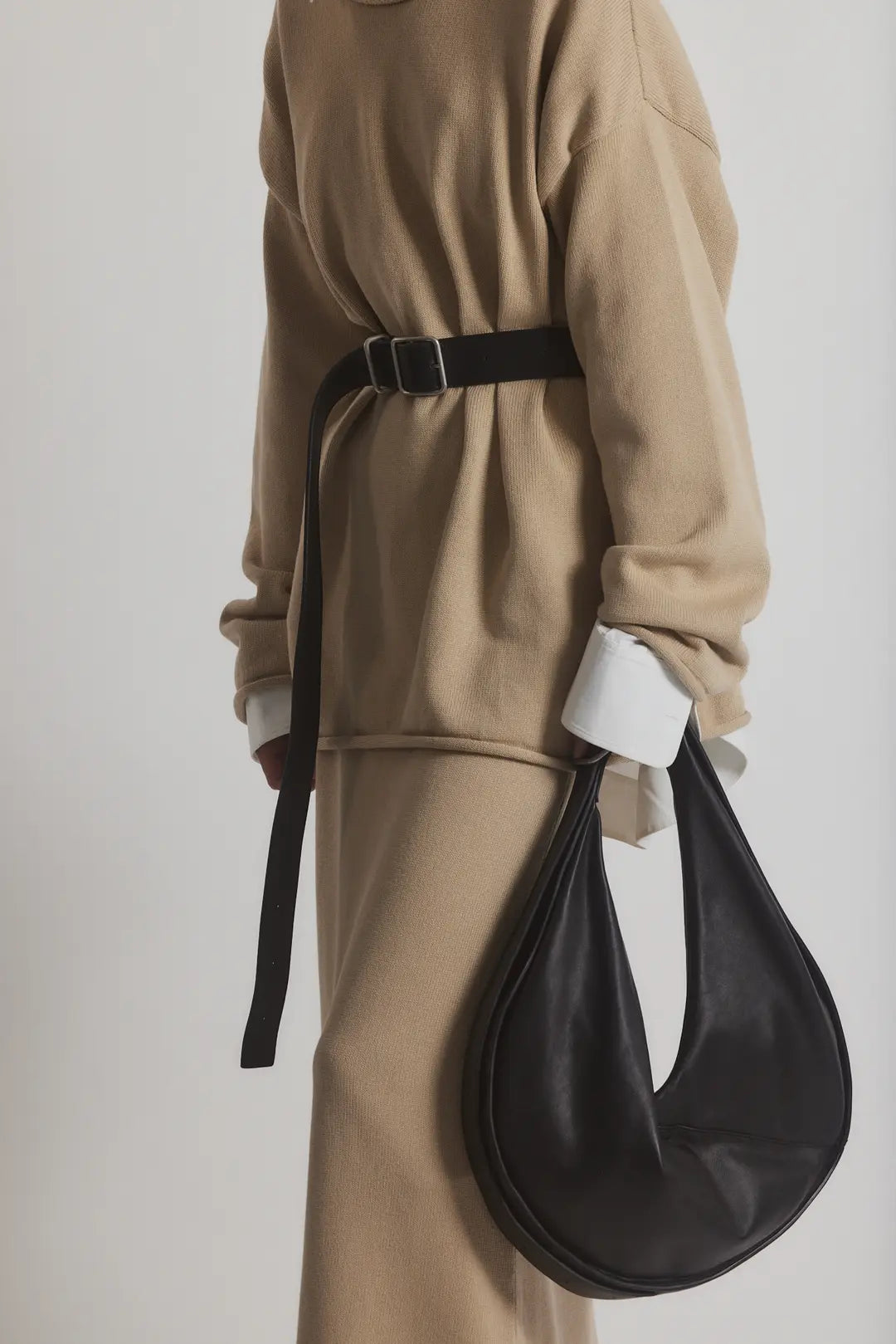 A person in a beige outfit, featuring the Bowe Belt by Janessa Leone with custom hardware, and white cuffs, wears a long skirt and holds a large bag made from black and earth brown regenerative leather, creating a modern and sophisticated look.