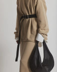 A person in a beige outfit, featuring the Bowe Belt by Janessa Leone with custom hardware, and white cuffs, wears a long skirt and holds a large bag made from black and earth brown regenerative leather, creating a modern and sophisticated look.