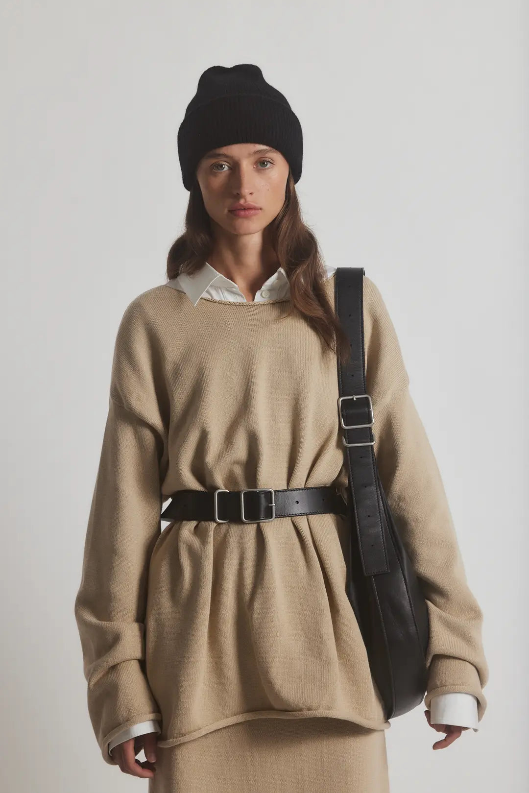 A person wears a black beanie, white shirt, and beige oversized sweater cinched with the Janessa Leone Bowe Belt. They carry a black shoulder bag made from locally crafted regenerative leather in Los Angeles and stand against a plain background.