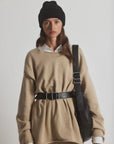 A person wears a black beanie, white shirt, and beige oversized sweater cinched with the Janessa Leone Bowe Belt. They carry a black shoulder bag made from locally crafted regenerative leather in Los Angeles and stand against a plain background.