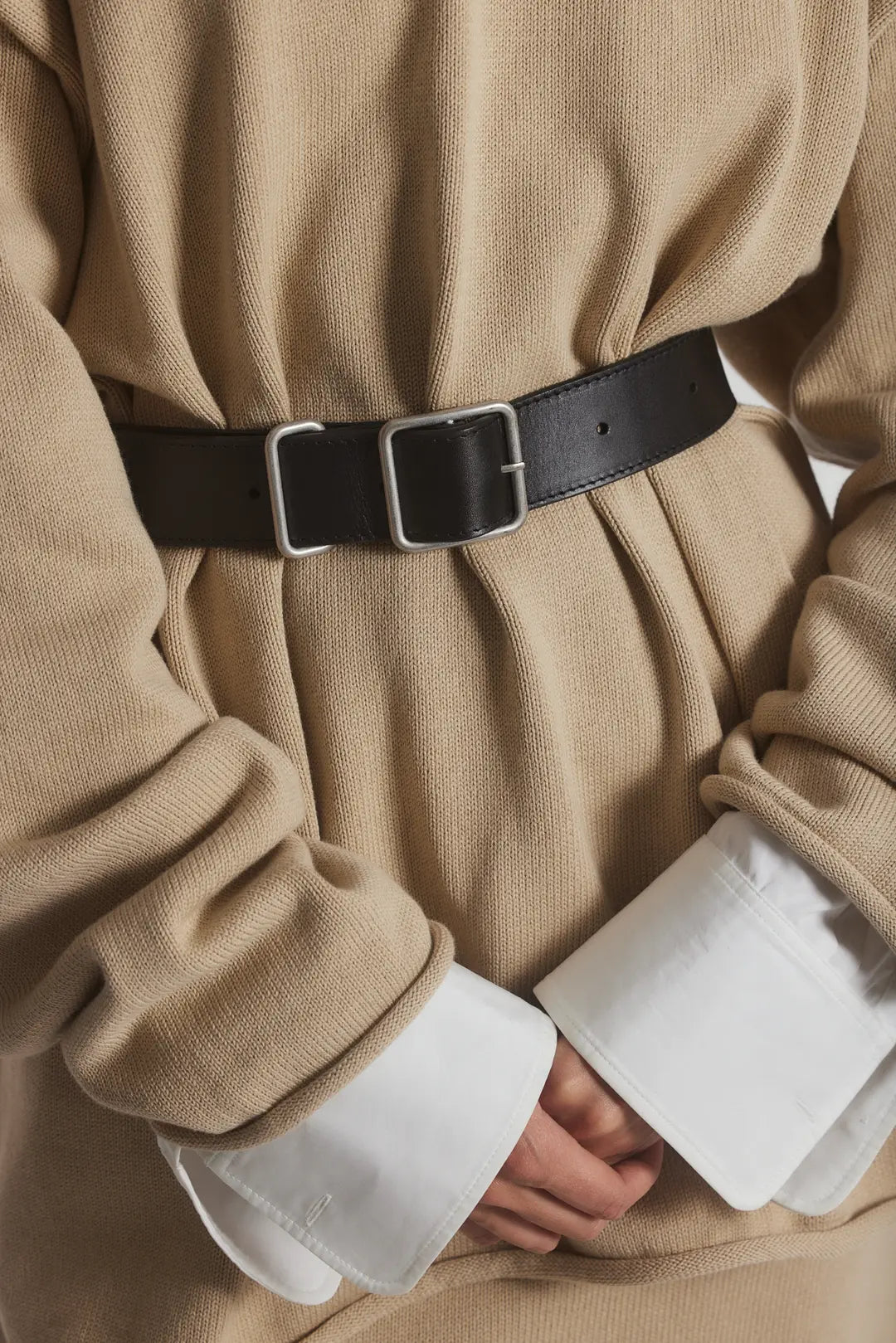 A person wears a beige sweater over a white shirt with wide cuffs, accessorizing with the Bowe Belt by Janessa Leone. The belt features custom-designed hardware and is crafted from black and earth brown regenerative leather. Their hands are clasped in front.