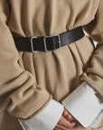 A person wears a beige sweater over a white shirt with wide cuffs, accessorizing with the Bowe Belt by Janessa Leone. The belt features custom-designed hardware and is crafted from black and earth brown regenerative leather. Their hands are clasped in front.