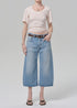 Wearing a short-sleeve, light-striped tee, Citizens Of Humanity/AGOLDE vintage culotte in sunlit indigo denim jeans, a brown belt, and black sandals, a person stands against a plain light gray background.