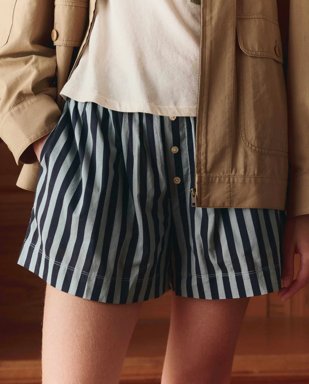 A person stands indoors wearing The Boxer Short by The Great Inc.—blue-and-white vertically striped cotton boxer shorts with buttons. The image is cropped to show only the lower torso and upper legs.