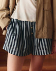 A person stands indoors wearing The Boxer Short by The Great Inc.—blue-and-white vertically striped cotton boxer shorts with buttons. The image is cropped to show only the lower torso and upper legs.