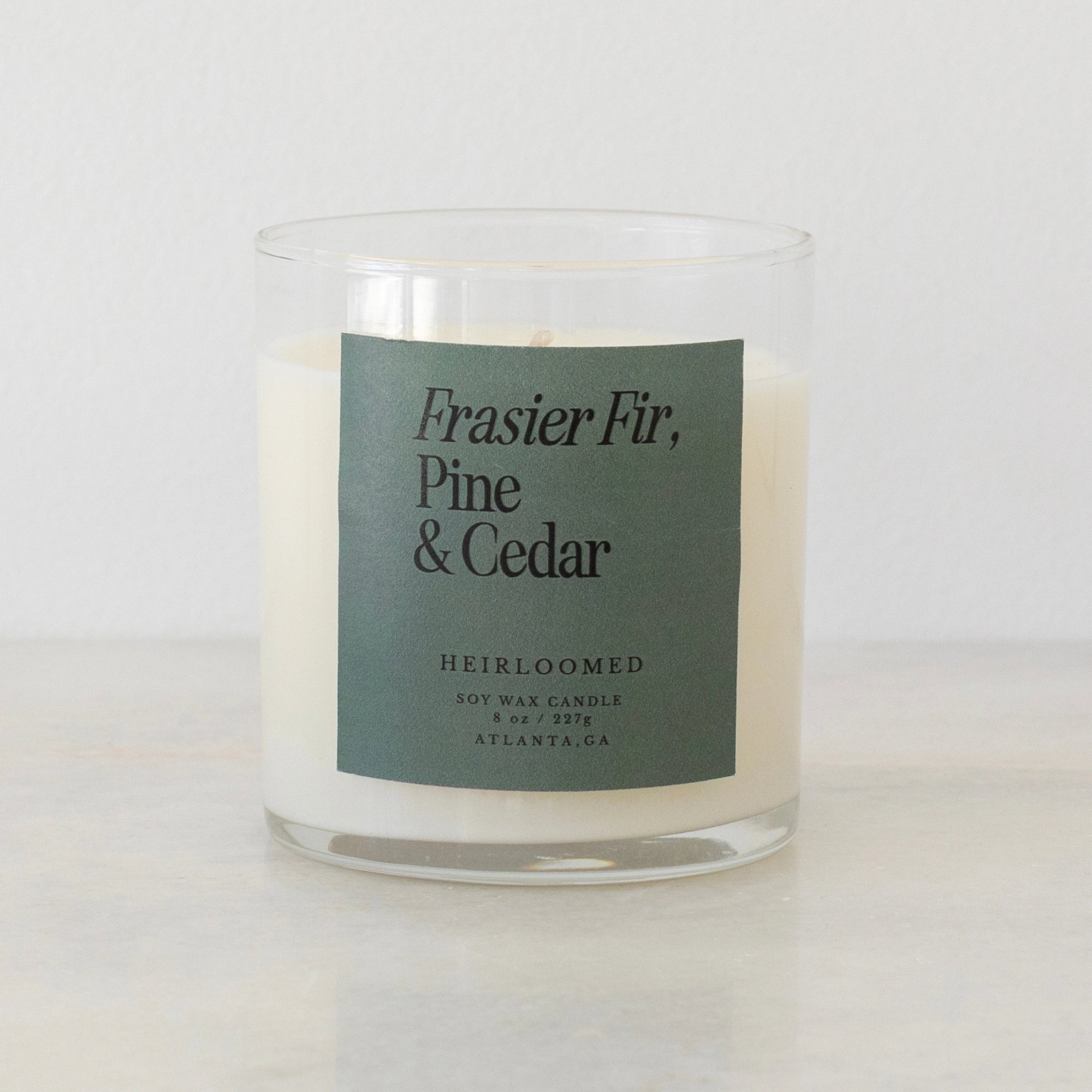 The Heirloomed Candle by Faire is a white soy wax candle in a clear glass jar with a green "Frasier Fir, Pine & Cedar" label. Poured in Atlanta, GA, it fills your space with festive Christmas tree scents.