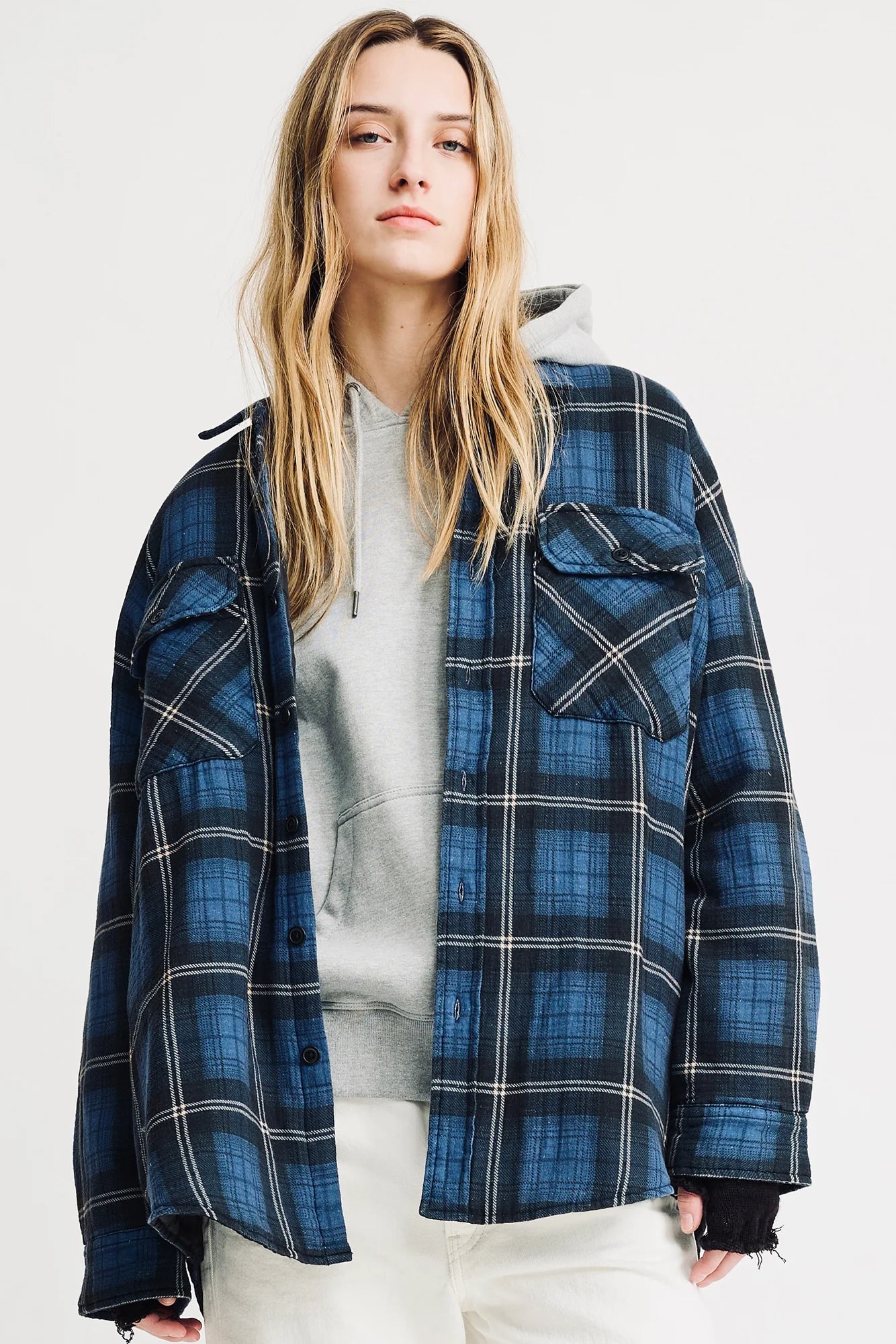 A person with long blonde hair wears a gray hoodie under the Denimist CPO SHIRT JACKET in blue and black plaid, paired with off-white cotton pants, standing against a plain light background.