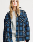 A person with long blonde hair wears a gray hoodie under the Denimist CPO SHIRT JACKET in blue and black plaid, paired with off-white cotton pants, standing against a plain light background.