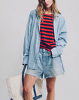 A woman in a light blue washed Maire Blue oversized button-down shirt, BROOKE SHORT MAIRE BLUE by Denimist with a raw hem, and holding a large white canvas tote bag with blue trim stands against a plain background.