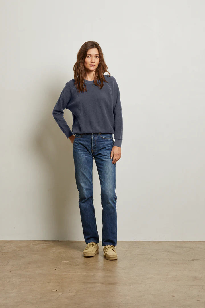 A woman with long brown hair stands against a plain wall, wearing the Perfectwhitetee Ziggy Reverse LS Sweatshirt in dark blue cotton fleece, blue jeans, and beige shoes. She has one hand in her pocket and looks relaxed.