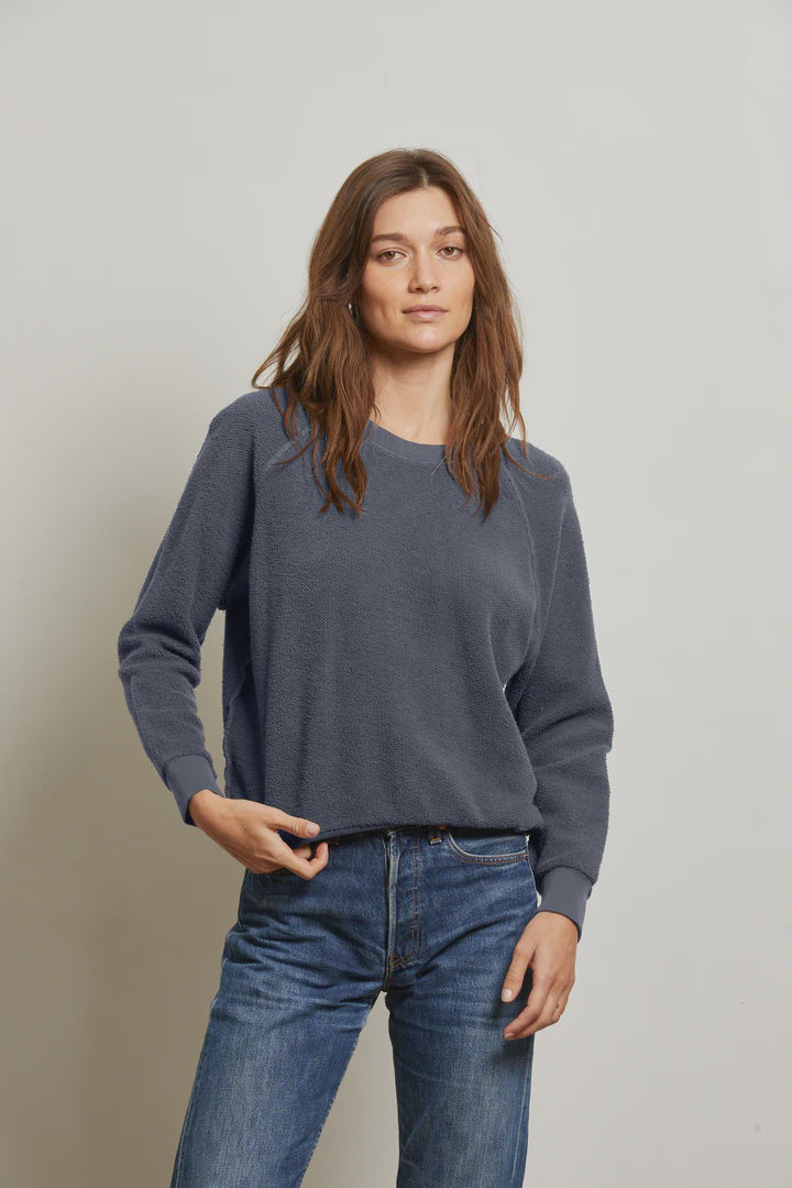 A woman with long brown hair wears the Perfectwhitetee Ziggy Reverse LS Sweatshirt and blue jeans, standing against a light gray background and looking at the camera with a neutral expression.