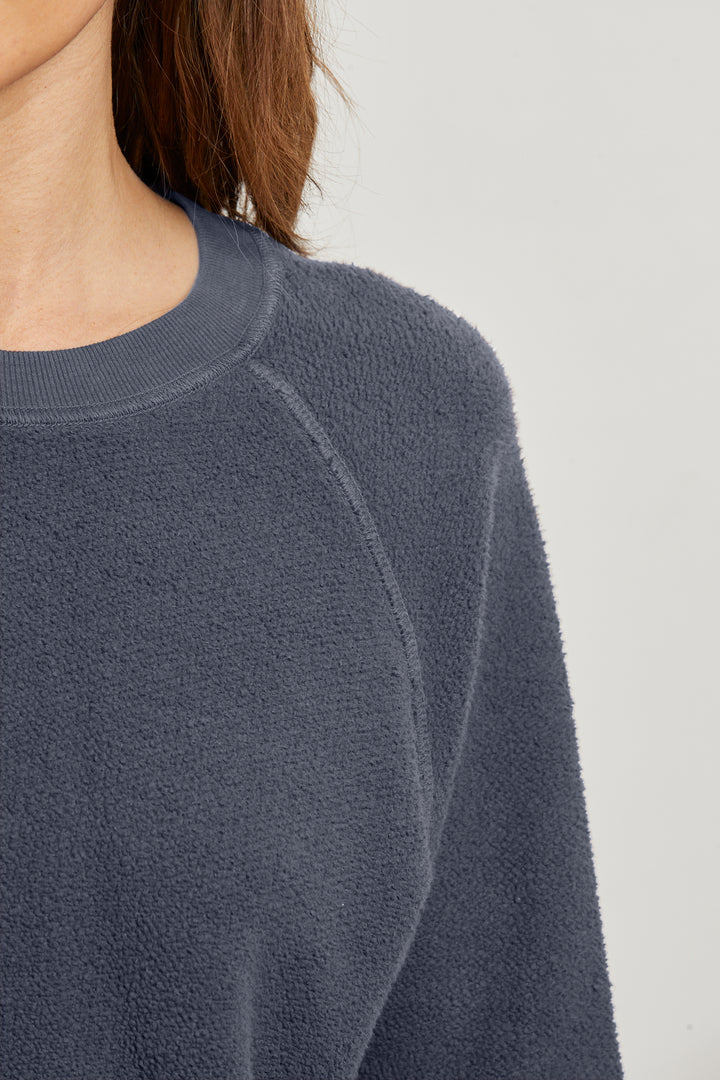 Close-up of a person wearing the Perfectwhitetee Ziggy Reverse LS Sweatshirt in textured dark blue cotton fleece, featuring a crew neckline and bracelet-length sleeves. Only their neck, shoulder, and upper chest are visible against a plain background.
