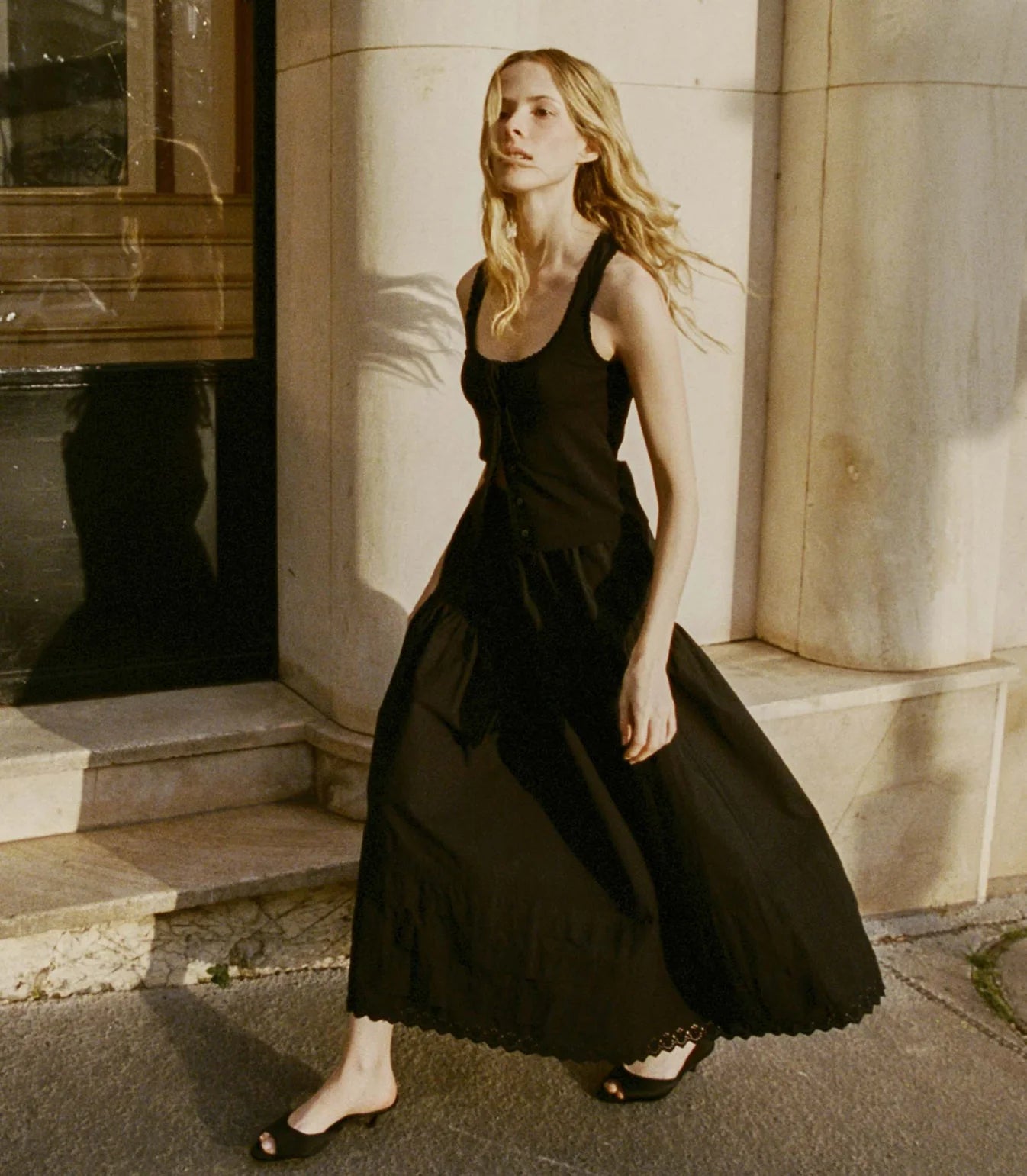 A woman with long blonde hair, wearing DOEN's Sebastiane Skirt as part of a sleeveless black dress and black sandals, walks on a sunlit sidewalk in front of a light-colored stone building.