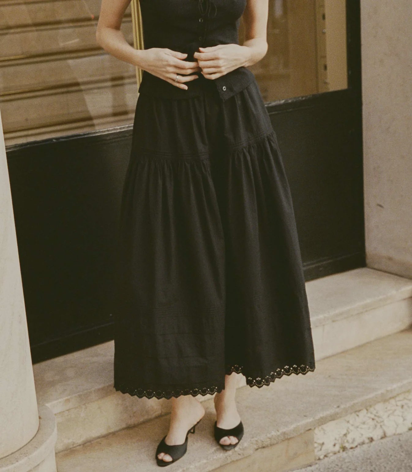 A person wearing the DOEN Sebastiane Skirt— a long black organic cotton skirt with scallop eyelet embroidery— and black open-toe shoes stands on a stone step before a building with a black door and pillar. Their upper face is not shown.