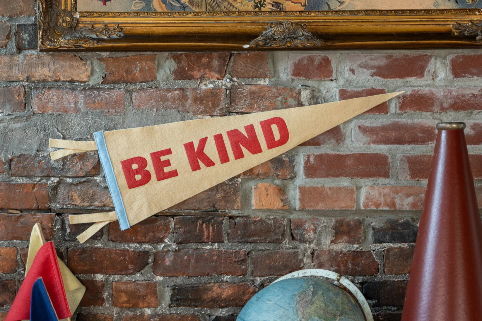 The BE Kind Pennant by Oxford Pennant, a bold red-lettered triangular flag, hangs on a brick wall. A globe, flags, and a brown cone-shaped object appear in the foreground, creating a positive atmosphere.