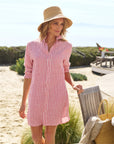A woman in the Frank & Eileen MARY Shirtdress S25 and a straw hat stands outdoors by a sandy beach, with greenery and a wooden deck behind her. A woven bag and towel sit on a nearby wooden chair.
