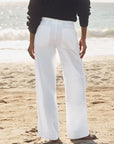 On a sandy beach with the sun reflecting off the ocean, a person faces the water wearing Frank & Eileen’s Limerick Carpenter Pant and a black long-sleeve top.