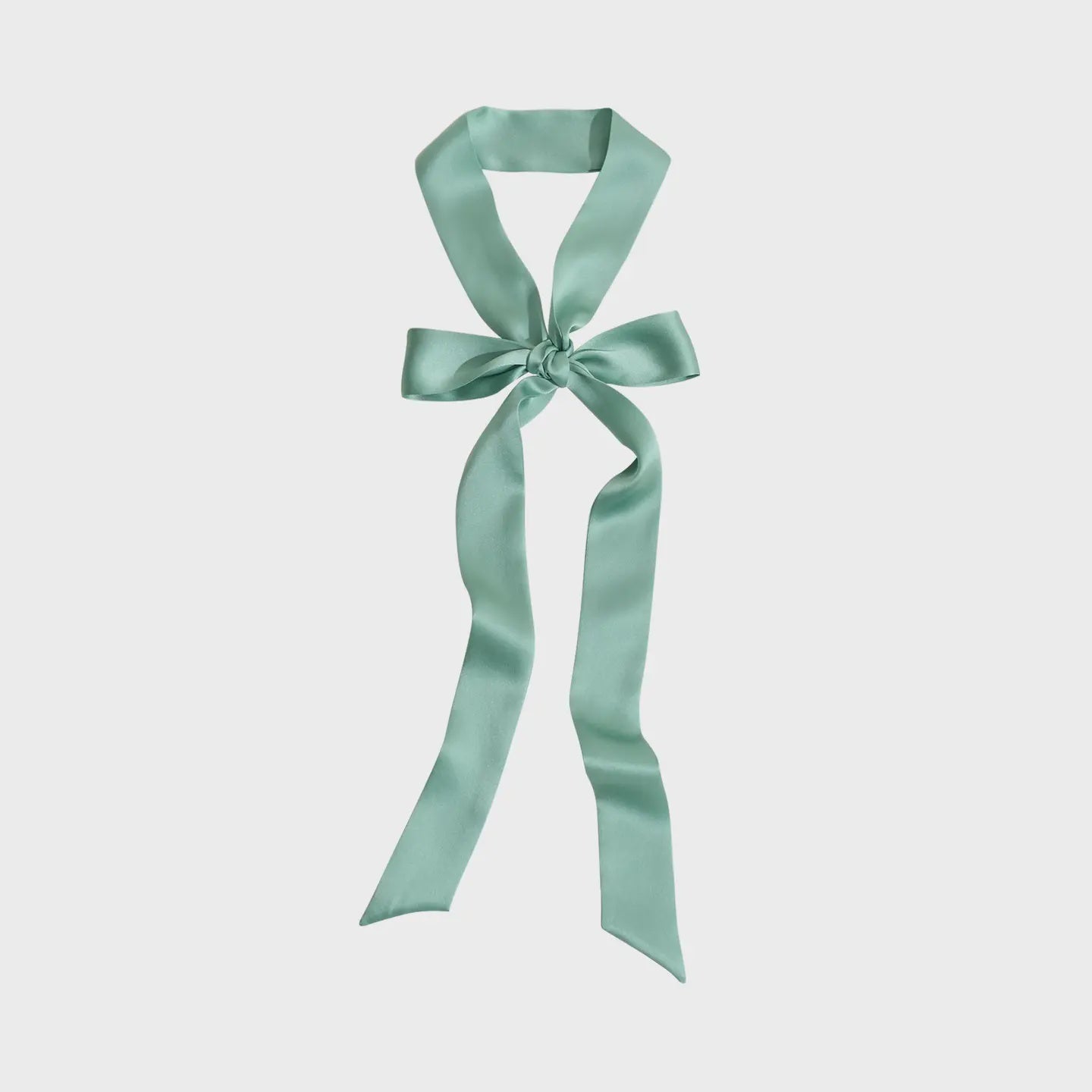 A vintage muse graces the scene with Faire's La Collectionneuse Silk Ribbon Scarf in Sage, tied into a decorative bow, its long, flowing tails elegantly draped against a plain white background.