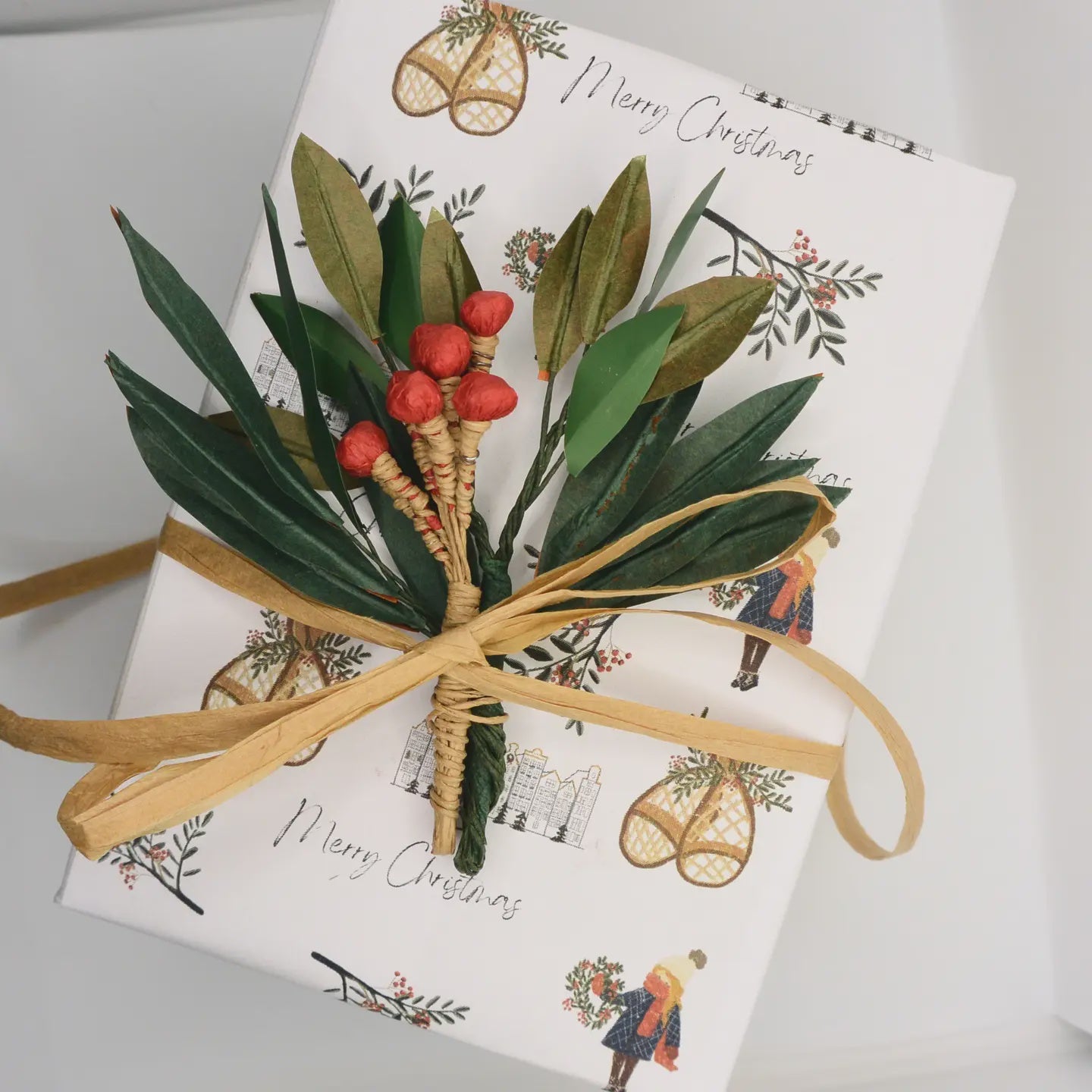 A Christmas gift wrapped in natural style paper adorned with festive illustrations and "Merry Christmas" is tied with a ribbon and features the Holly Floral Christmas Present Topper by Faire, showcasing a handmade arrangement of green leaves and red berries that embodies charming Christmas decor.