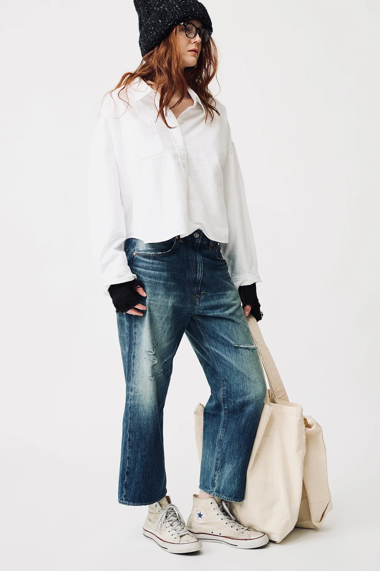 A person wearing a loose white shirt, Denimist HAISLEY SLOUCH BF JEAN in blue, white Converse sneakers, black beanie, and glasses stands holding a large beige tote bag against a plain white background.