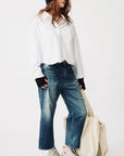 A person wearing a loose white shirt, Denimist HAISLEY SLOUCH BF JEAN in blue, white Converse sneakers, black beanie, and glasses stands holding a large beige tote bag against a plain white background.
