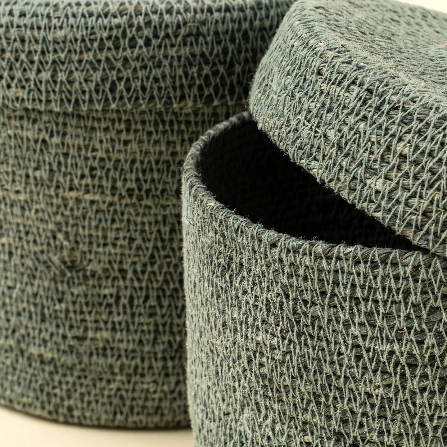 Close-up of two Tajine Seagrass Baskets by Indaba, featuring textured green woven designs. One basket’s lid is partially open to reveal a dark interior, while the other stands behind—ideal for stylish Moroccan-inspired storage.