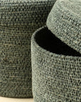Close-up of two Tajine Seagrass Baskets by Indaba, featuring textured green woven designs. One basket’s lid is partially open to reveal a dark interior, while the other stands behind—ideal for stylish Moroccan-inspired storage.